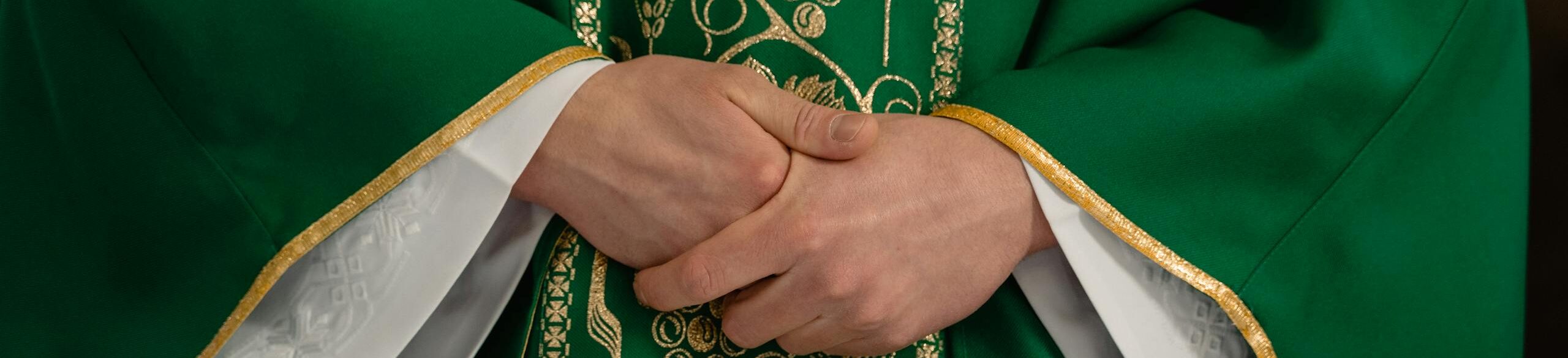 A close-up view of a priest's hands clasped in prayer wearing a green chasuble with gold embroidery.